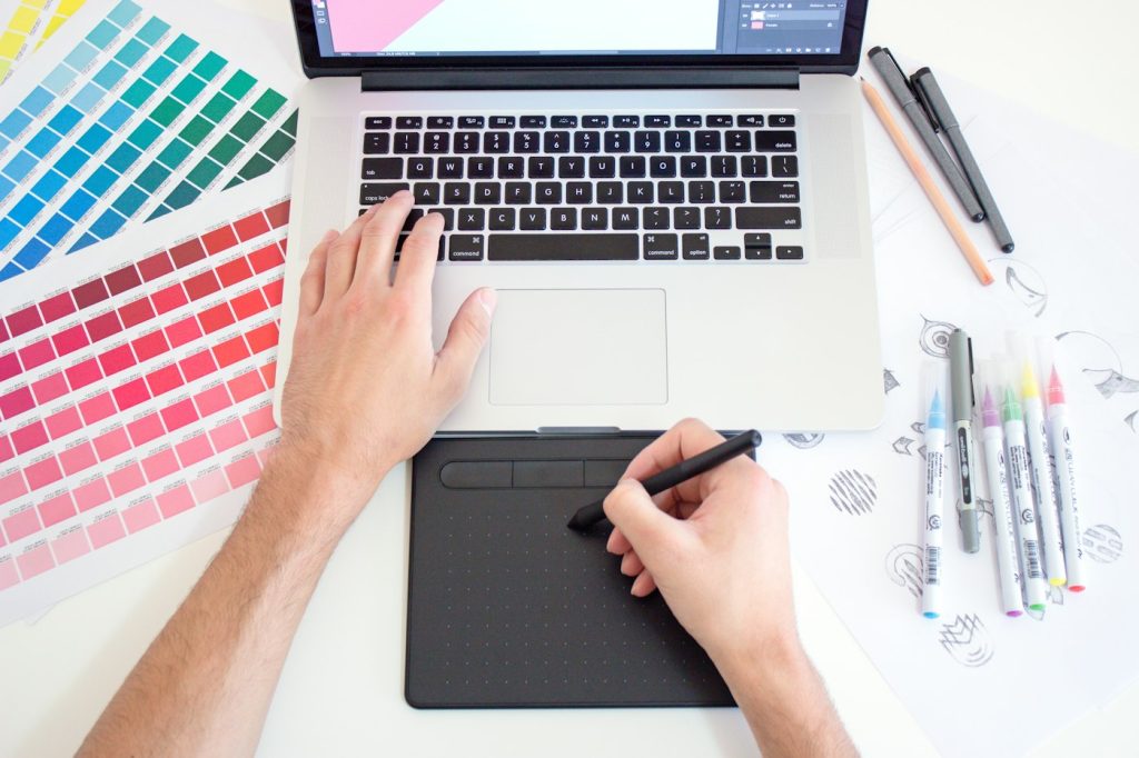 Designer working on a laptop and drawing tablet with colour swatches and markers spread across the desk